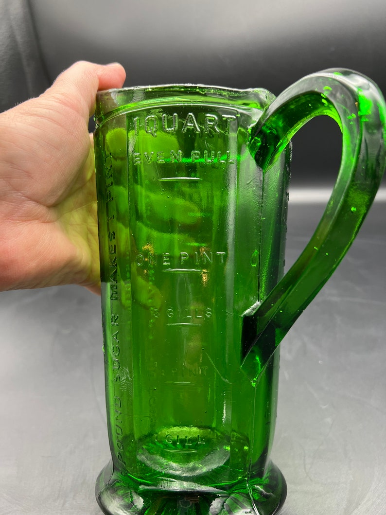 May include: A vintage green glass measuring pitcher with a large handle. The pitcher has measurement markings for quarts, pints, and gills. The pitcher is made of thick, durable glass and has a classic design. The pitcher is a kitchenware item.