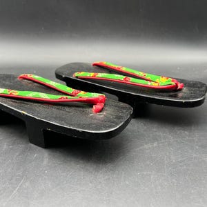 May include: Pair of traditional Japanese geta sandals. The wooden platform sandals are black with green and red fabric straps. The straps have a floral pattern. The geta are elevated by two wooden blocks.