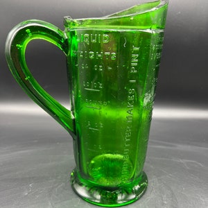 May include: A vintage green glass measuring pitcher with a handle. The pitcher has embossed measurement markings for liquid weights, including ounces and pints. The pitcher is made of clear green glass.