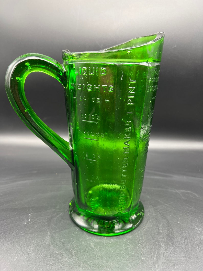 May include: A vintage green glass measuring pitcher with a handle. The pitcher has embossed measurement markings for liquid weights, including ounces and pints. The pitcher is made of clear green glass.