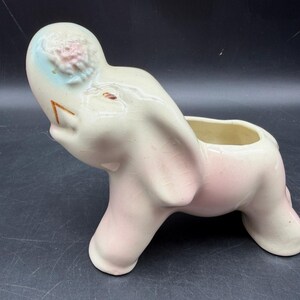 May include: A ceramic elephant planter in cream and pink. The elephant has a raised trunk, a light blue patch on its head with a pink floral design, and a rectangular opening on its back for planting. The planter is a decorative item.