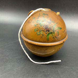May include: A round, brown container with a floral design in green, purple, and white. A white cord is attached to the top. The container is made of two connected halves.