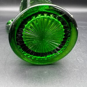 May include: A green glass pitcher, viewed from the bottom, showcasing a detailed, textured design. The base features a circular pattern with radiating lines and a dark, contrasting border. The pitcher's body curves upwards, with a handle visible on the side.