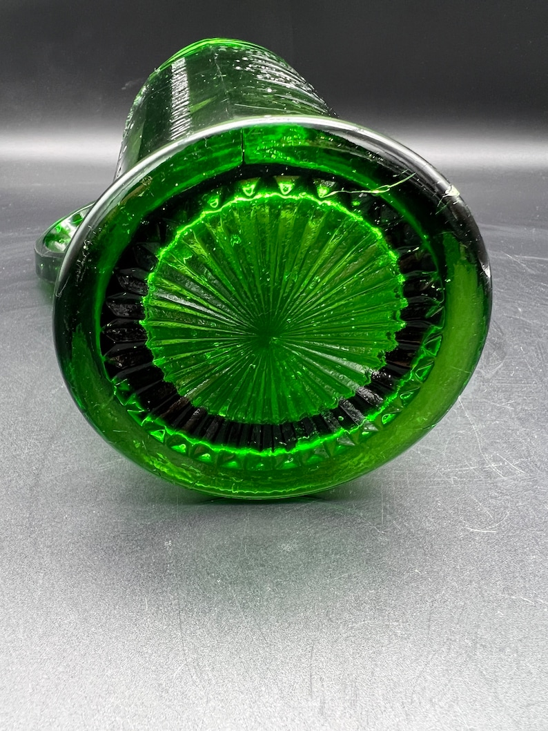 May include: A green glass pitcher, viewed from the bottom, showcasing a detailed, textured design. The base features a circular pattern with radiating lines and a dark, contrasting border. The pitcher's body curves upwards, with a handle visible on the side.