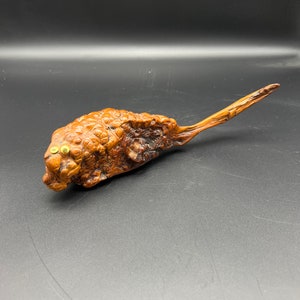May include: A unique, hand-carved wooden sculpture with a textured, bulbous body and a long, pointed tail. The sculpture features two small, round, golden eyes. The wood has a rich, natural brown color, with variations in tone and texture.
