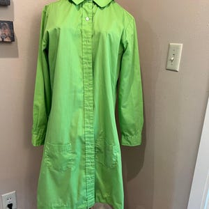 May include: A bright green, long-sleeved coat with a rounded collar and two front pockets. The coat is buttoned up the front and is displayed on a wicker dress form. The coat is a solid, vibrant color.