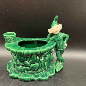 May include: A vintage green ceramic planter shaped like a well, with a smiling elf figure leaning against it. The well has a textured stone appearance and a small bucket detail. The elf is wearing a green outfit and hat.