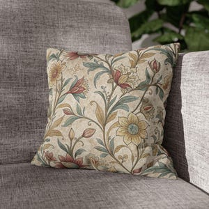 May include: Square decorative pillow with a floral pattern. The pillow features a beige background with a repeating design of flowers and leaves in shades of red, yellow, and green. The pillow is placed on a gray sofa.