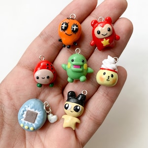 Puede incluir: Colección de colgantes de arcilla polimérica hechos a mano. Los colgantes presentan varios personajes de dibujos animados en colores brillantes como naranja, rojo, verde y amarillo. Cada colgante tiene un lazo plateado para sujetarlo a un llavero o collar.
