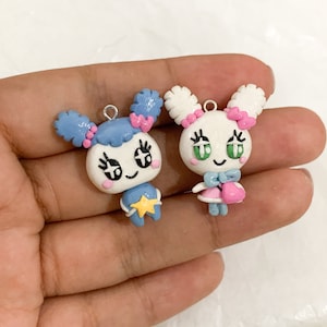 Puede incluir: Dos colgantes hechos a mano, estilo dibujos animados. Uno es azul con una estrella, el otro es blanco con un lazo rosa y un pequeño bolso rosa. Ambos tienen ojos negros, mejillas rosas y un lazo plateado para colgar.