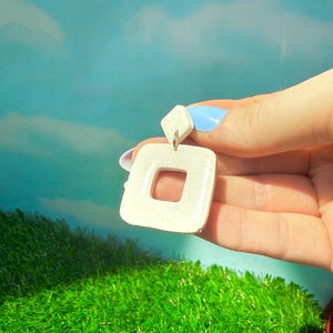 May include: Cream-colored square earrings with a smaller square stud. The earrings have a glossy finish and a square cut-out in the center. The earrings are held against a backdrop of a blue sky and green grass.