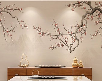 Op de afbeelding: Een modern woonkamerinterieur met een lichtbeige muur met een bloemige muurschildering van een bloeiende kersenboom. De kamer heeft een houten salontafel met een glazen blad, een houten kast met laden en een moderne kroonluchter.
