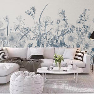 Op de afbeelding: Een witte hoekbank met grijze kussens en een grijs plaid staat voor een muur met een blauw en wit bloemenbehang. Een witte salontafel met een witte poef staat voor de bank.