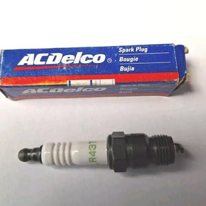 May include: An ACDelco spark plug with a white ceramic insulator and a black metal threaded base. The white insulator features the green text R43T. It is accompanied by its original blue, red, and white rectangular cardboard packaging box.