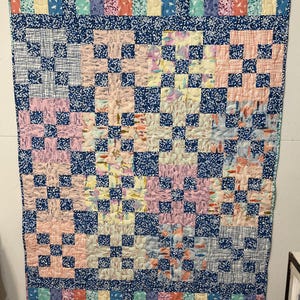 May include: A handmade quilt with a patchwork design. The quilt features squares of various patterns and colors, including pink, blue, yellow, and patterned fabrics. The border and some squares are a dark blue with white speckles. The top border has vertical stripes.