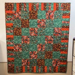 May include: A square patchwork quilt featuring a repeating grid pattern of fabric squares. The design alternates between solid green textured fabric, a floral print with red and orange flowers, and a dark print featuring small orange jack-o'-lanterns.