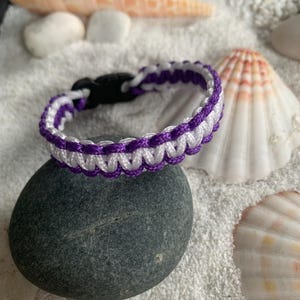 Peut inclure: Un bracelet tressé violet et blanc avec une fermeture noire. Le bracelet est sur une surface blanche avec des coquillages.