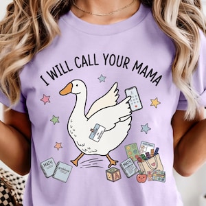 May include: A light purple t-shirt featuring a white goose illustration holding a smartphone. The text reads I WILL CALL YOUR MAMA. Surrounding the goose are colourful stars, maths notebooks, a tote bag, an apple, and alphabet blocks.