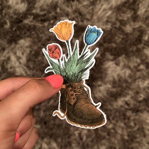 May include: A brown leather boot with a bouquet of three tulips in it. The tulips are red, yellow, and blue.
