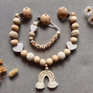Könnte beinhalten: Ein Holz-Beißring mit einem Regenbogen-Anhänger und eine separate Holzperlenkette mit dem Namen "Mathea" darauf. Der Beißring und die Kette bestehen beide aus natürlichen Holzperlen und haben weiße herzförmige Perlen.