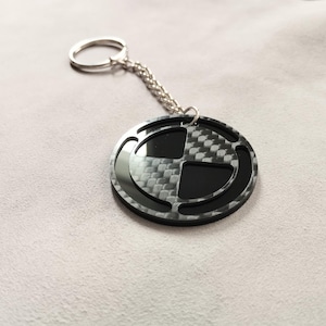 May include: A circular keychain featuring a black and gray carbon fiber pattern with a cut-out design. The round pendant is attached to a silver-toned metal split ring by a short chain. The surface of the pendant has a glossy, reflective finish.