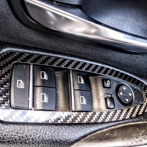 Puede incluir: Primer plano de un panel de puerta de coche con botones de control de ventanas y espejos. Los botones son negros con iconos blancos, en un marco de estilo fibra de carbono. El panel está rodeado de cuero negro.