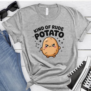 May include: A gray t-shirt featuring the text "KIND OF RUDE POTATO" above a cartoon potato character with an angry expression. The potato is light brown with pink cheeks and small arms and legs. The shirt is paired with blue jeans and black sunglasses.