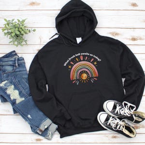 May include: Black hooded sweatshirt with a colorful rainbow and sun graphic. The text "What fresh hell awaits us today?" is printed above the rainbow. The sweatshirt is paired with blue jeans and black canvas sneakers.