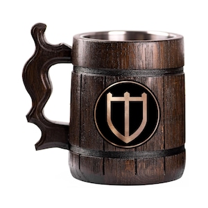 May include: A dark brown wooden beer mug with a decorative handle and a stainless steel interior. The mug features a black emblem with a gold shield and a stylized letter "T". The mug is designed to hold beverages.