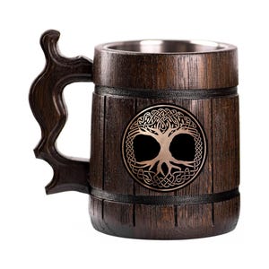 May include: A dark brown wooden beer mug with a decorative handle. The mug features a metal interior and a bronze-colored emblem of a tree of life. The mug is designed with vertical wooden planks and black bands.