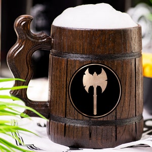 May include: A wooden beer mug filled with beer and foam. The mug has a dark brown finish with a decorative handle. A gold axe emblem is on the front. The mug is sitting on a white and black patterned cloth.