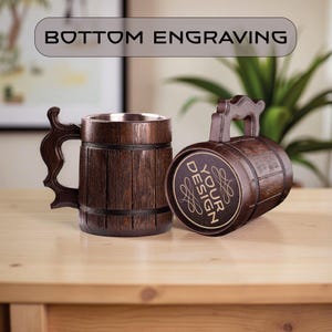 May include: Two wooden beer mugs with a barrel design. One mug is upright, the other is on its side. The mugs have a dark brown finish with a silver interior. The text "BOTTOM ENGRAVING" is above the mugs, and "YOUR DESIGN" is on the side of one mug.