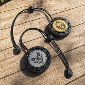 Op de afbeelding: Twee zwarte ronde accessoires met verstelbare koorden. Eén heeft een schedelontwerp op een gouden achtergrond, de andere een lachend clowngezicht. Beide hebben een getextureerde, donkergrijze rand en zwarte koorden met verstelbare toggles.