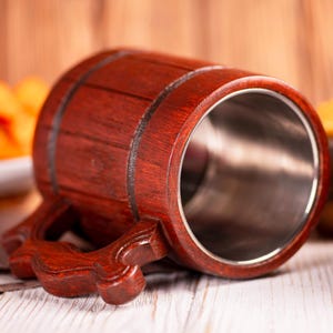 May include: A wooden beer mug with a dark reddish-brown finish. The mug has a stainless steel interior and a decorative handle. The mug is made of wood and has a barrel-like shape.