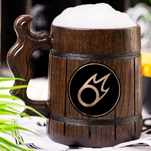 May include: A wooden beer stein filled with beer and a thick layer of foam. The stein has a dark brown finish and a decorative handle. A black circle with a gold design is on the front of the stein. The stein is sitting on a white and black patterned cloth.