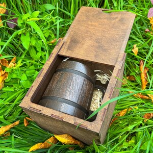 May include: A wooden gift box with an open lid, revealing a miniature barrel and two small glass jars. The barrel is dark brown with black bands. The box is set in a bed of green grass and fallen leaves.