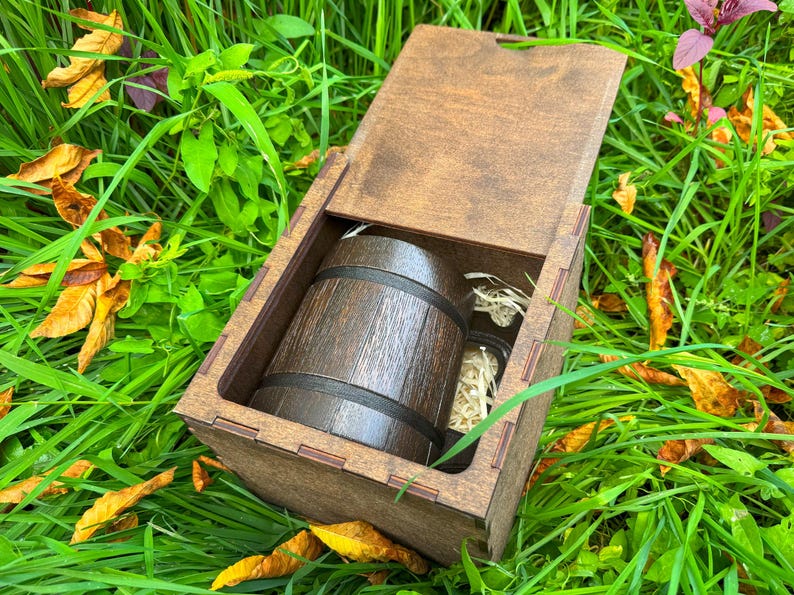 May include: A wooden gift box with an open lid, revealing a miniature barrel and two small glass jars. The barrel is dark brown with black bands. The box is set in a bed of green grass and fallen leaves.