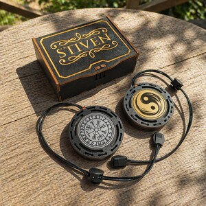 Peut inclure: Une boîte en bois noire avec l'inscription dorée "STIVEN" est posée à côté de deux pendentifs ronds. L'un des pendentifs présente un motif de boussole, l'autre un symbole yin-yang. Les pendentifs sont reliés à des cordons noirs.