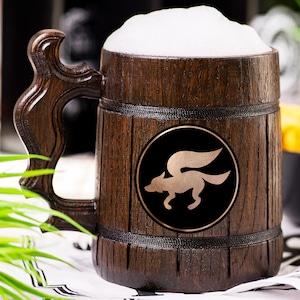 May include: A wooden beer mug filled with beer and foam. The mug has a dark brown finish with a decorative handle. A circular emblem with a winged fox is on the front. The mug is sitting on a white and black patterned cloth.