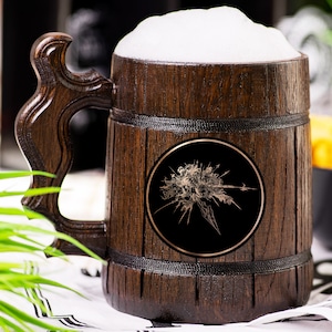 Può includere: Un boccale di birra in legno pieno di birra e schiuma. Il boccale ha una finitura marrone scuro con un manico decorativo. Un emblema circolare nero con un design dorato è sulla parte anteriore. Il boccale è su un panno bianco e nero.