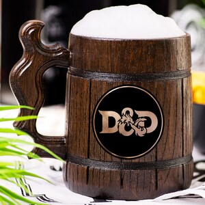 May include: A wooden beer tankard filled with beer and foam. The tankard has a dark brown finish with a carved handle. A black circle with the letters "D&D" in copper-coloured lettering is on the front. The tankard is on a white surface.