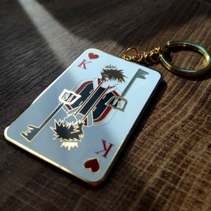 Puede incluir: Un llavero plateado con un diseño de carta de juego que presenta una ilustración de personaje. La carta es el Rey de Corazones, con un borde dorado y un llavero dorado.