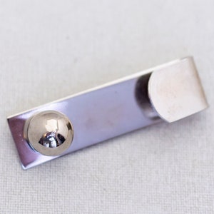 May include: A silver-toned metal money clip with a rectangular body and a rounded end. A small, shiny sphere is attached to the front. The clip is designed to hold folded bills securely.