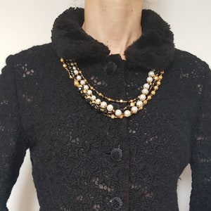 May include: Black lace jacket with a faux fur collar and three buttons. The jacket features a floral pattern and is paired with a multi-strand pearl necklace. The bottom of the jacket has a scalloped edge. The jacket is worn over a black and white patterned skirt.