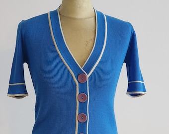 May include: A blue short-sleeved cardigan with a white trim and five purple buttons.