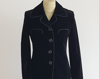 May include: A black velvet skirt suit displayed on a mannequin. The suit features a blazer with a notched collar, three buttons, and faux pocket details. The skirt is a simple, knee-length design. Black piping trims the edges of the jacket.