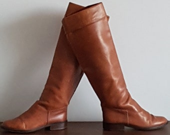 May include: A pair of knee-high brown leather boots. The boots have a classic design with a slight heel and a smooth, polished finish. The boots are angled, showcasing their shape and texture against a neutral background.