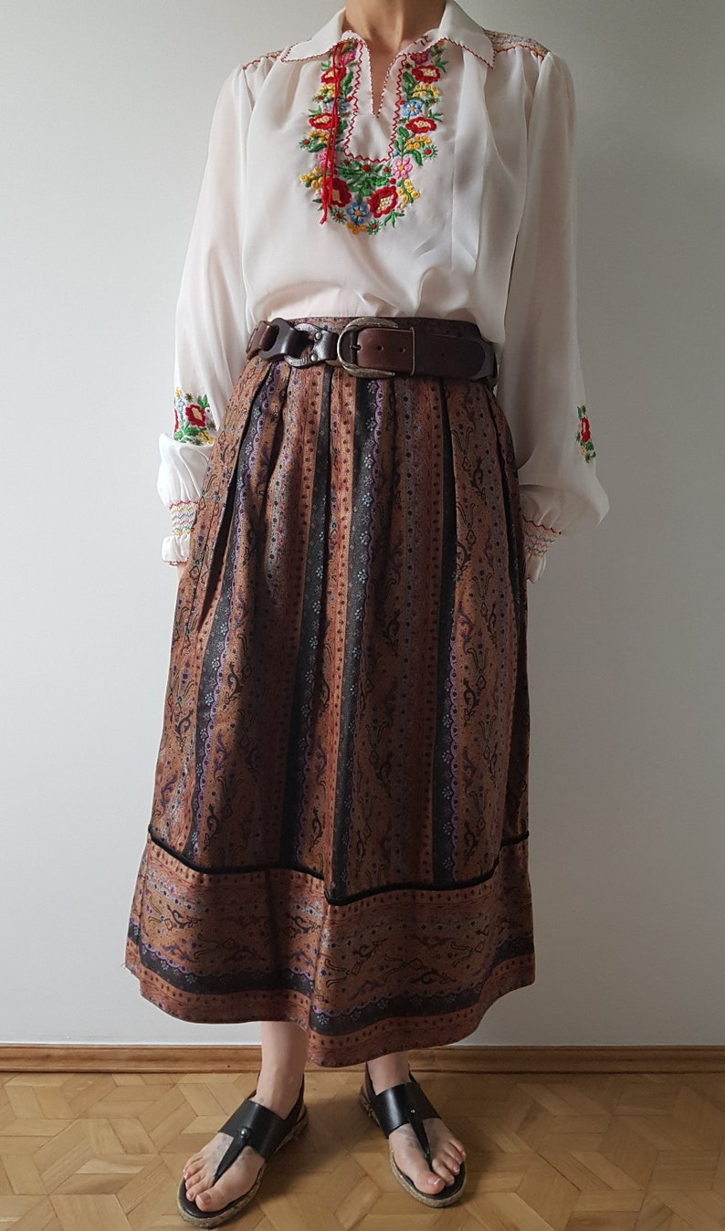 May include: A long brown skirt with a black and purple floral pattern. The skirt has a wide waistband with a brown leather belt. The skirt is paired with a white blouse with colorful embroidered flowers.