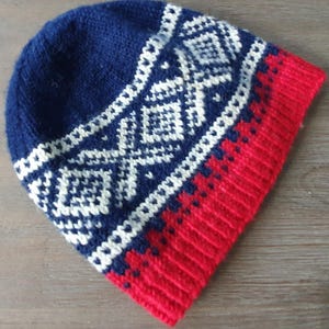 Może przedstawiać: Dzianinowa czapka beanie w kolorze granatowym, czerwonym i białym. Czapka ma geometryczny wzór w kolorze białym na granatowej części, z jednolitym czerwonym paskiem na dole. Czapka jest wykonana z miękkiego, ciepłego materiału.