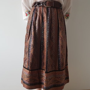May include: A brown and black patterned skirt with a black velvet trim. The skirt has a high waist and is pleated. It is worn with a white blouse with embroidered flowers.